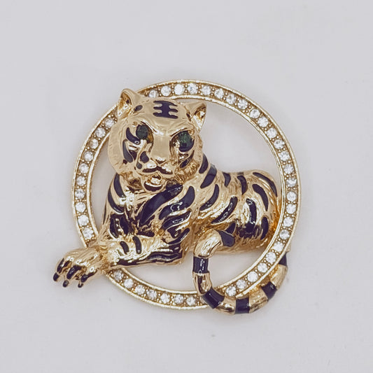 A vintage brass brooch in the shape of a tiger with a gold finish, featuring a vibrant green crystal eye and a circle of white crystals around it.