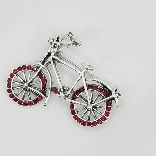 A brooch shaped like a bicycle with red crystals on the wheel spokes and silver-colored alloy frame.