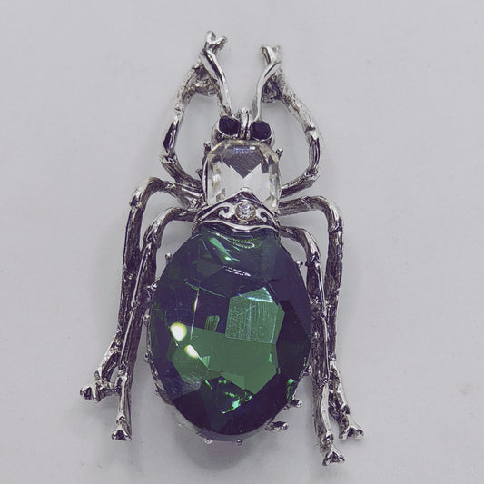 A silver-plated brooch in the shape of a beetle, adorned with a large green crystal rhinestone and small clear crystals.