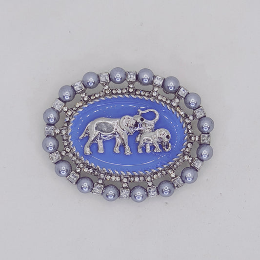 A brooch depicting two elephants, one adult and one baby, with the adult elephant wearing a crown. The brooch is primarily blue with silver accents, designed to resemble a pearl enamel.