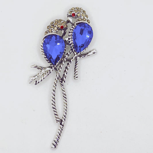 A brooch depicting two blue crystal birds perched on a silver branch.