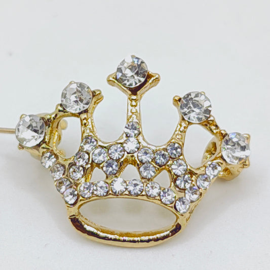 A gold tone crown brooch adorned with white stones, featuring a safety pin clasp.
