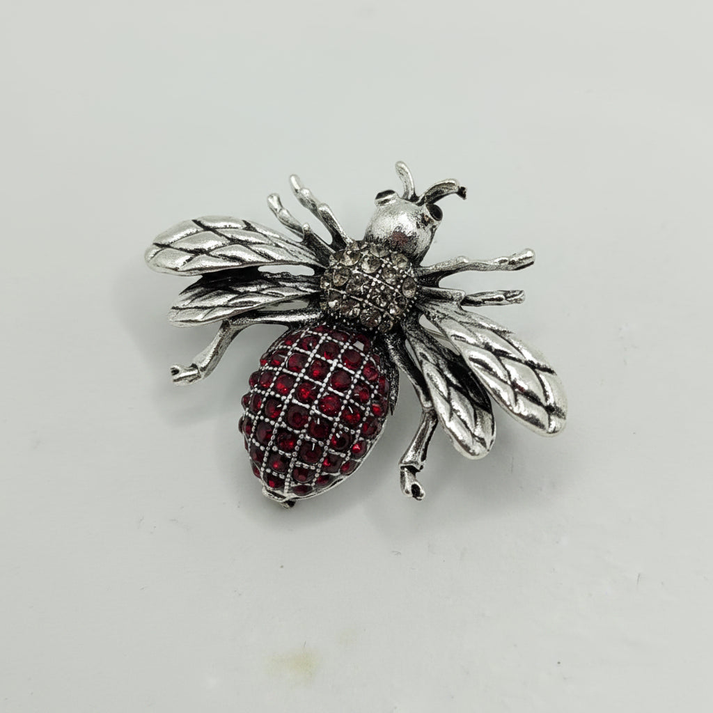 Silver bee-shaped brooch with red gemstones on a light gray background