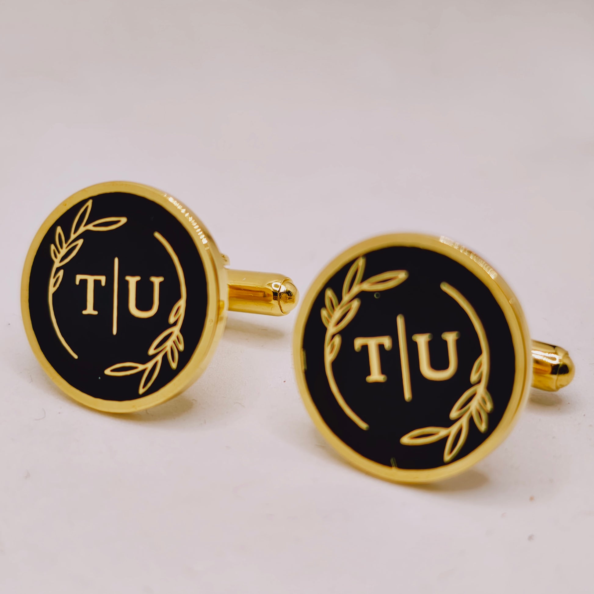 Gold and black cufflinks with 'T/U' initials on a white background